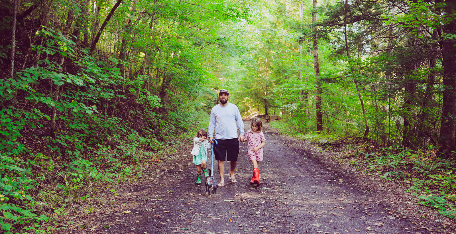 Man, father, with two young children, girls age 7 and 4 take a walk together with the family dog on a leash in a nature area, candid and happy